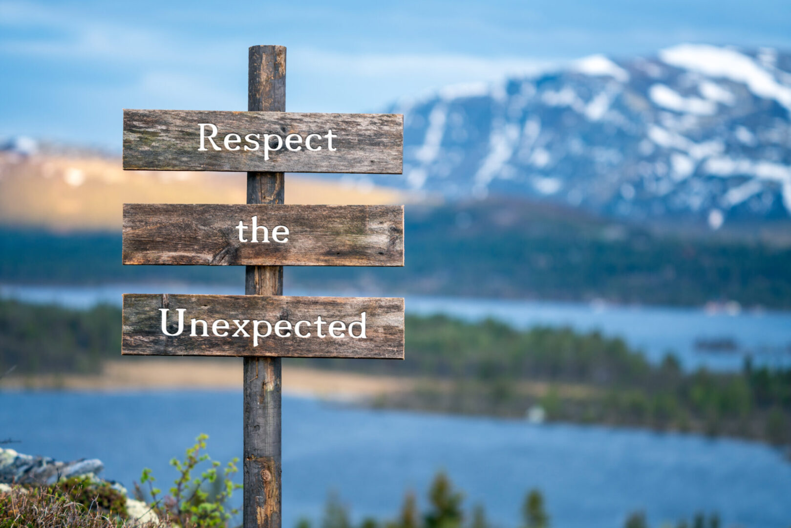 respect the unexpected text on wooden signpost outdoors in landscape scenery during blue hour. Sunset light, lake and snow capped mountains in the back.